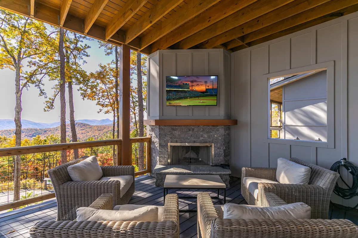 A cozy, open living area features a comfortable seating arrangement positioned around a modern stone fireplace. Walls of windows frame breathtaking mountain views, while a large TV is integrated into the space for movie nights. The vaulted ceiling opens up the room and bathes it in natural light, creating a welcoming atmosphere for gatherings.