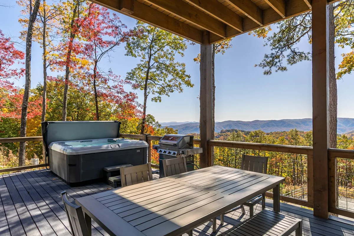 A spacious deck with a private hot tub and outdoor dining table, framed by a mountain view and vibrant autumn foliage. The hot tub sits to one side, steam visibly rising, inviting relaxation, while the dining area is set for alfresco meals. The deck railings and grill add to the sense of a well-equipped, multifunctional outdoor living area.