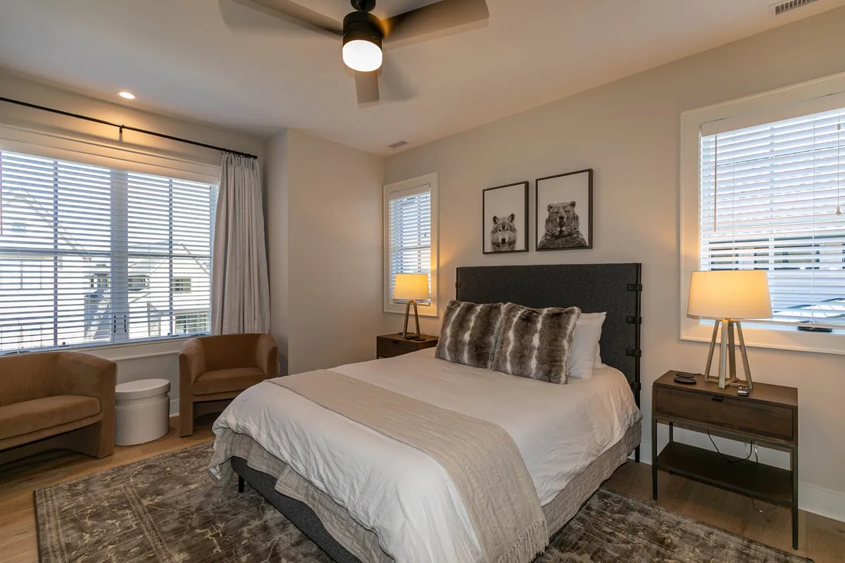 A serene bedroom featuring a bed with plush pillows, inviting artwork above the headboard, bedside tables with warm lighting, and comfortable chairs set beside large windows that allow natural light to gently illuminate the space. The overall atmosphere is calm and cozy, ideal for unwinding at the end of the day.