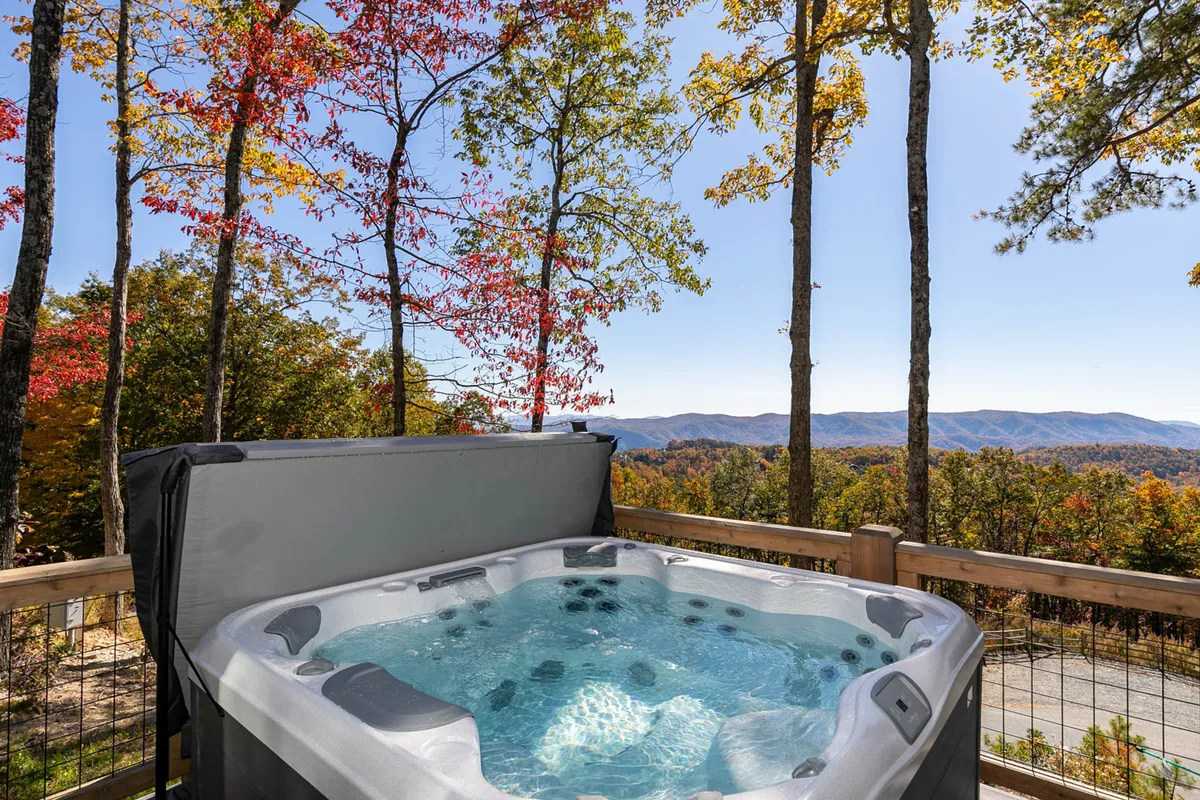 A bubbling hot tub sits at the edge of a deck overlooking mountains ablaze with fall colors. The scene is tranquil and secluded, emphasizing the experience of soaking in warm water while immersed in nature. The sunset light and surrounding foliage add to the sense of seasonal enjoyment and privacy.