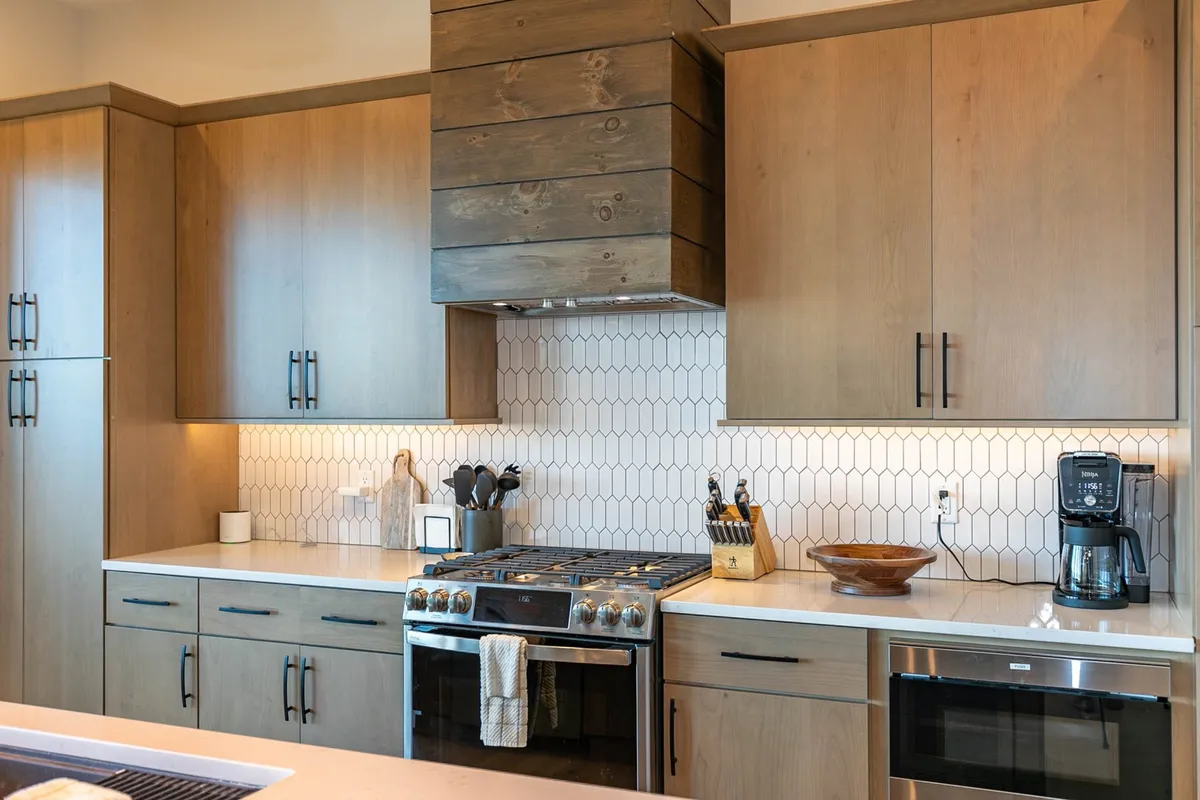 A warmly lit kitchen interior featuring a central island, stainless steel stove with a wooden range hood, and a striking hexagonal backsplash. The kitchen is designed for hosting and cooking, with ample counter space and high-end appliances displayed, inviting both meal preparation and social gatherings.