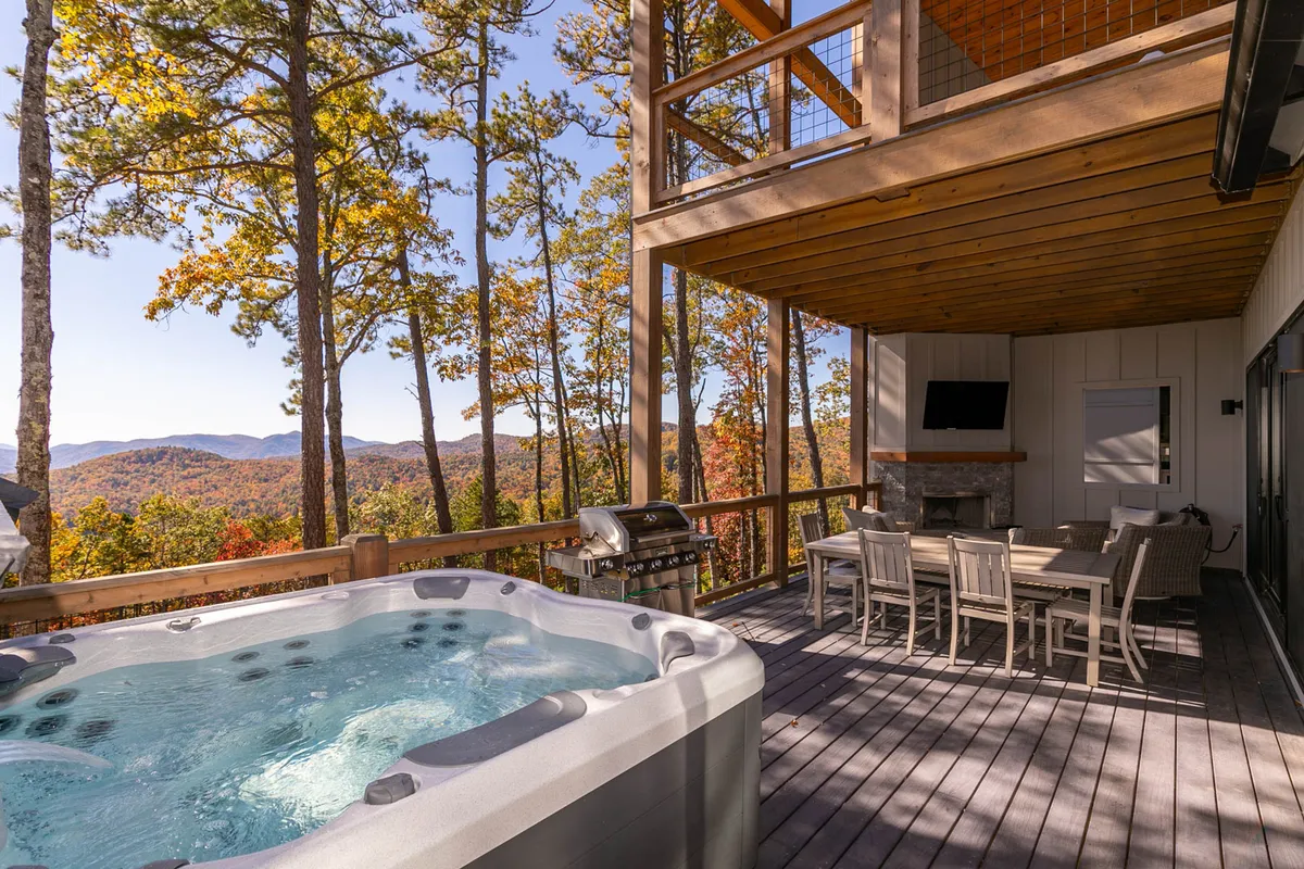 This exterior image features a private hot tub on a deck with sweeping mountain views, an outdoor dining table, grill, and plenty of space for relaxing under the stars. The setting is serene and ideal for romantic evenings or celebratory dinners in nature.
