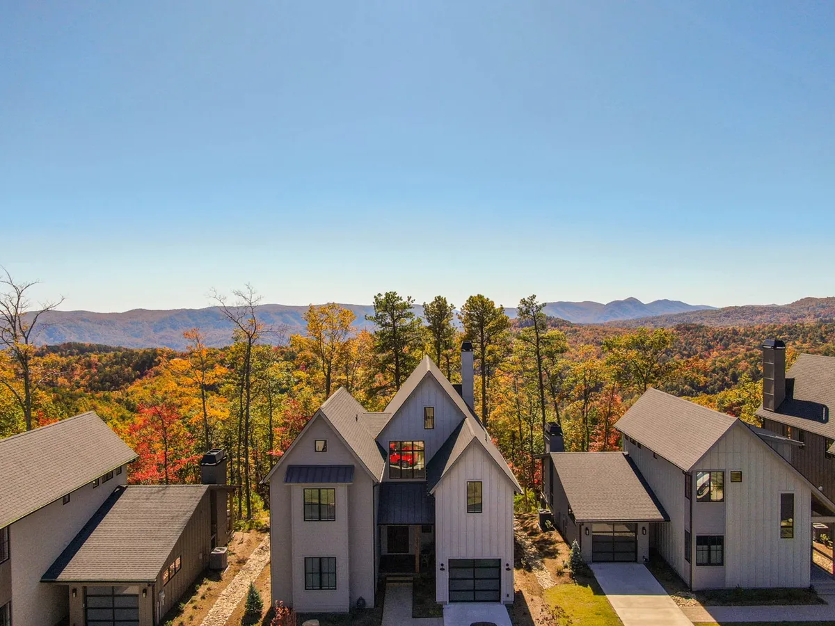 The image showcases a striking modern mountain home framed by vibrant autumn foliage, set against a breathtaking backdrop of rolling Blue Ridge Mountains. The expansive deck invites relaxation as the sun sets over the peaks, hinting at outdoor gatherings and serene moments in nature's embrace.