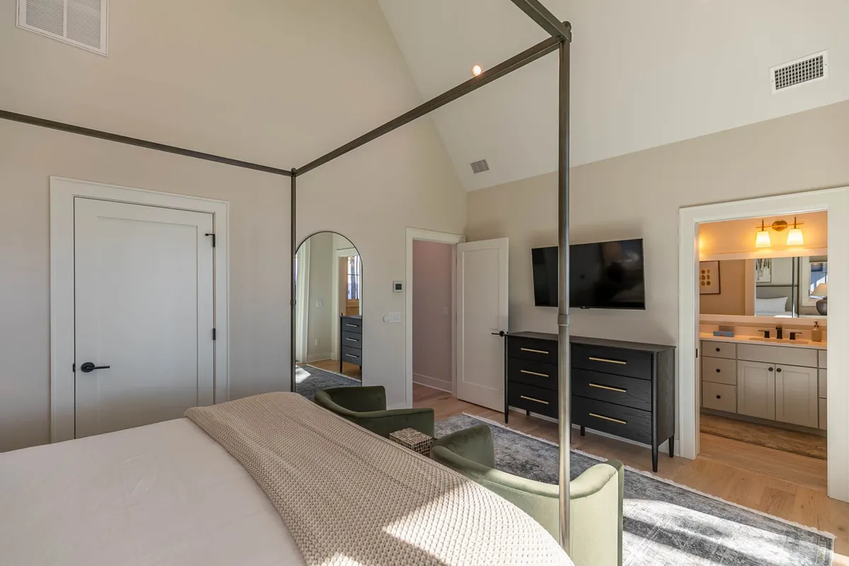 A spacious bedroom showcasing a four-poster bed dressed with plush bedding, green accent chairs for reading or relaxing, a large mirror, a sleek black dresser, and a TV. Sunlight streams into the room, enhancing the inviting and peaceful atmosphere.